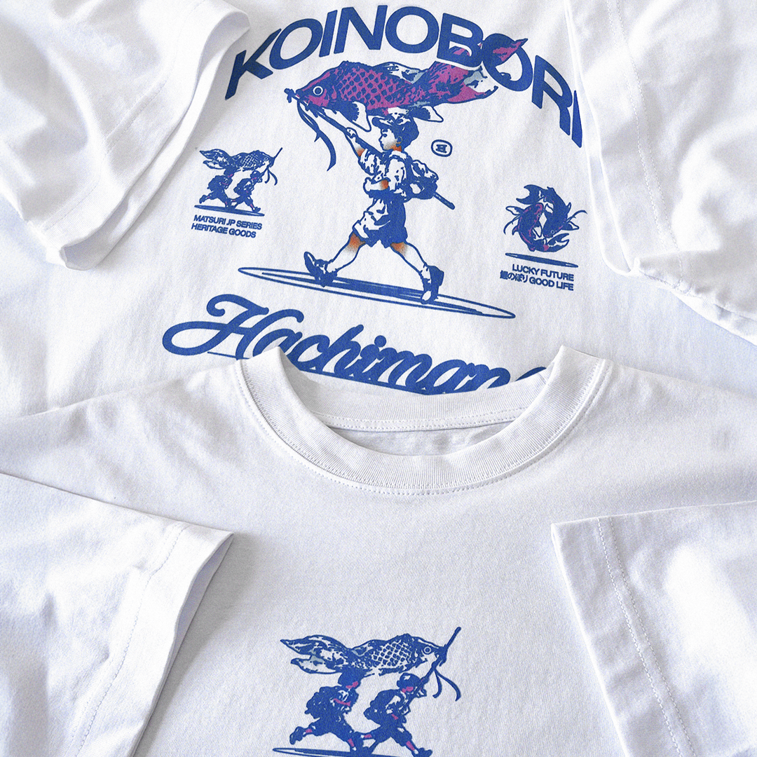White graphic t-shirt featuring blue and pink printed designs with Japanese text and a surfer character, shown in a flat-lay arrangement.